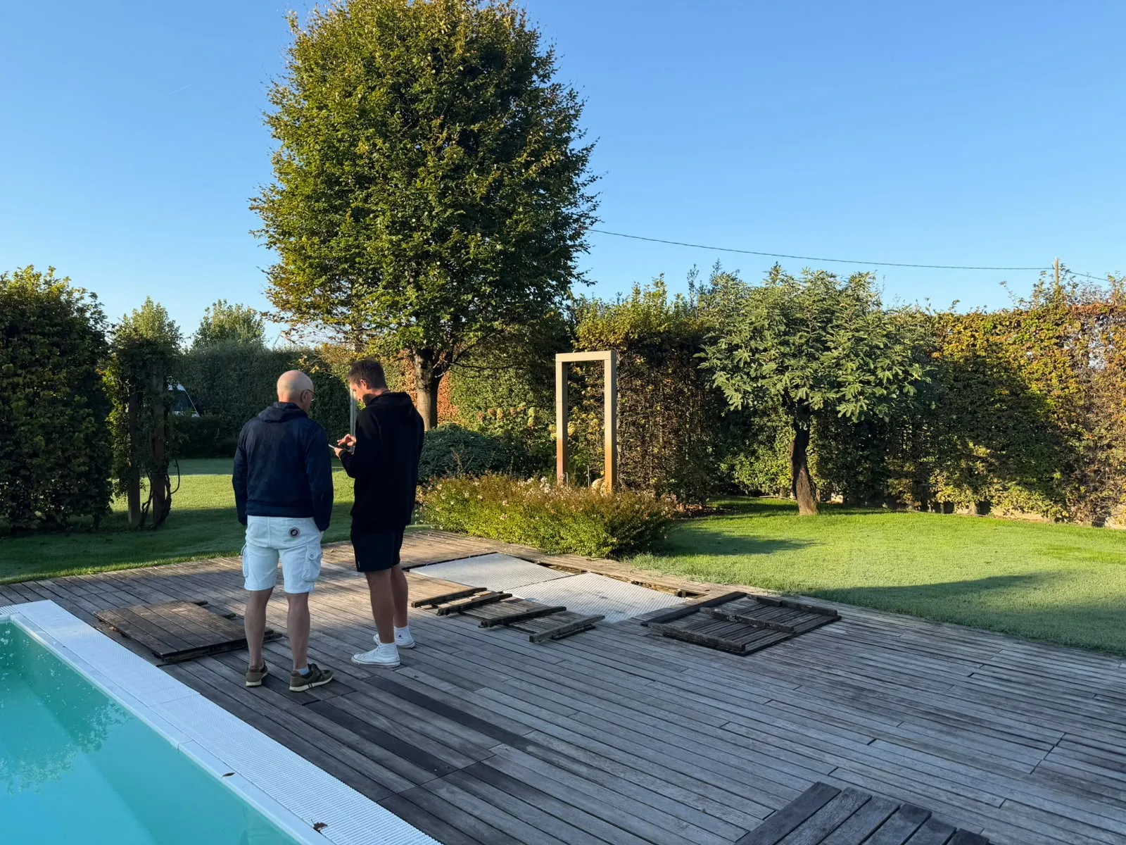 Due persone sul bordo di legno della piscina guardano insieme un telefono, con il prato, le siepi e un grande albero dietro